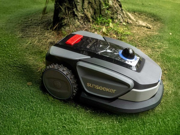 Sunseeker S4 robot lawn mower on a green lawn beside a tree trunk with dry grass and fallen leaves around its roots