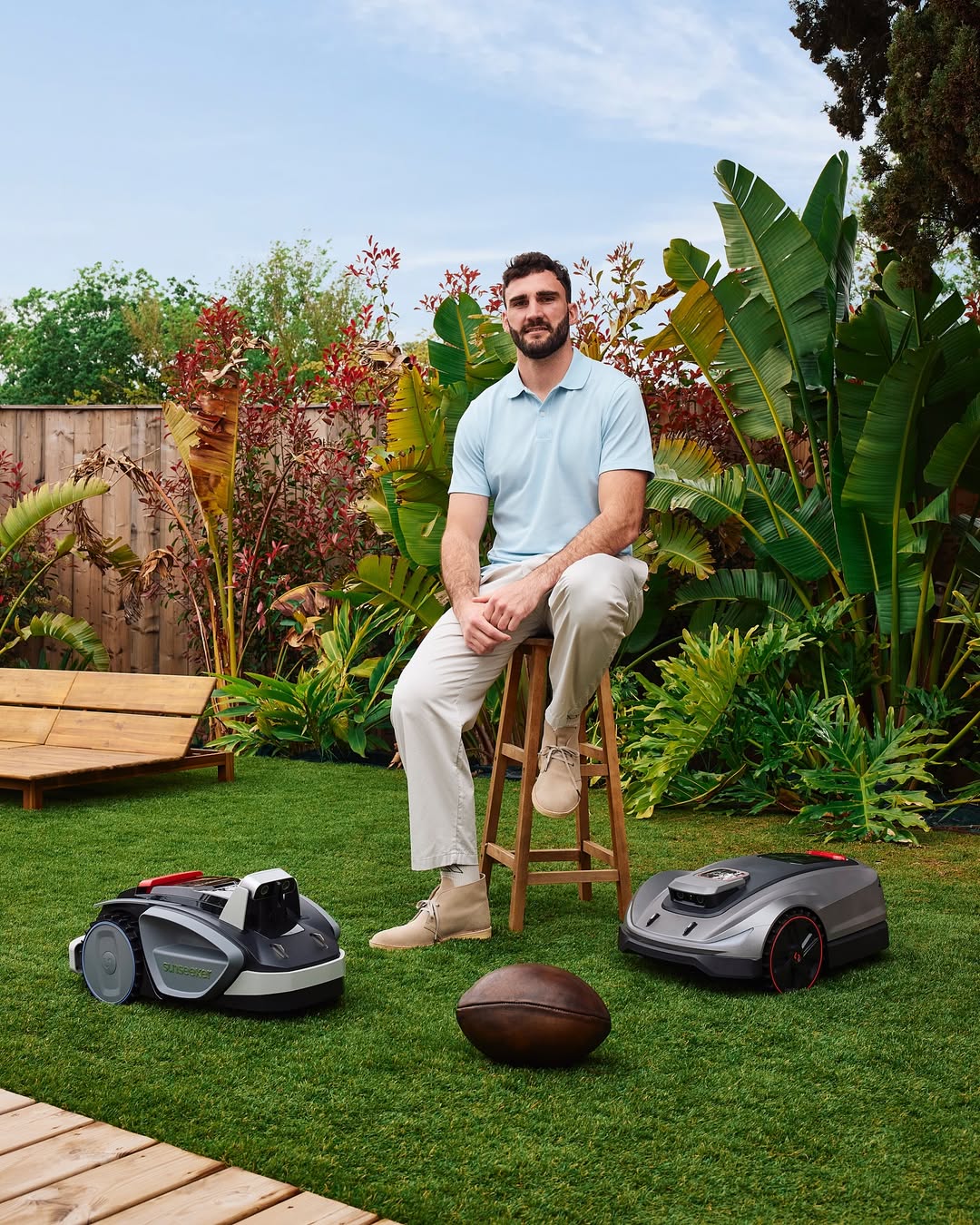 Man sitting on a stool in a garden with two robotic lawn mowers and a football on the grass.