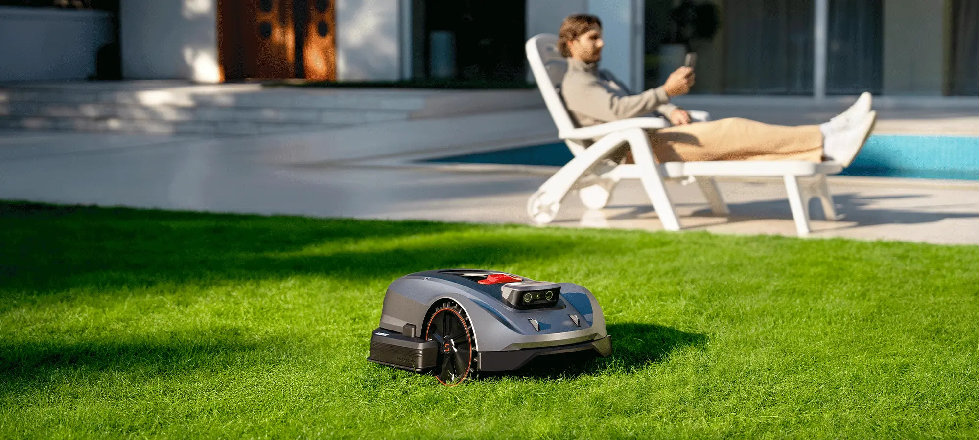 Robotic lawn mower on grass with a person using a smartphone in the background